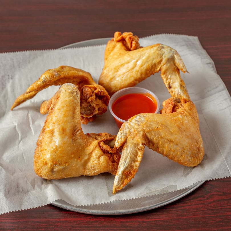 Four crispy fried chicken wings and a small cup of red dipping sauce on parchment paper.