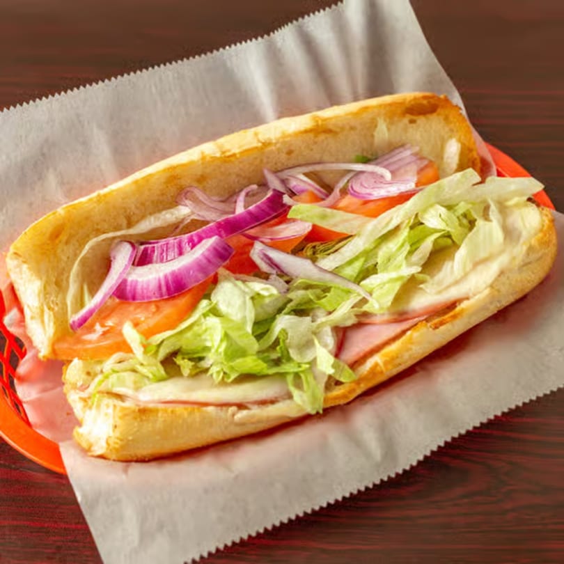 A delicious submarine sandwich with ham, cheese, lettuce, tomatoes, and red onions in a red basket.
