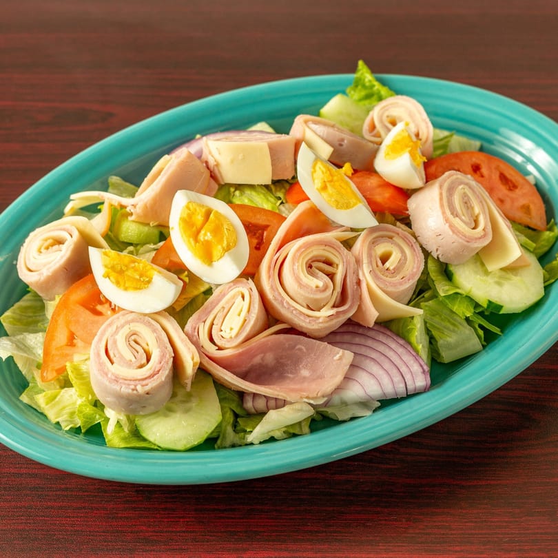 Chef salad with ham, cheese, hard-boiled eggs, tomatoes, cucumbers, and red onion on a teal plate.